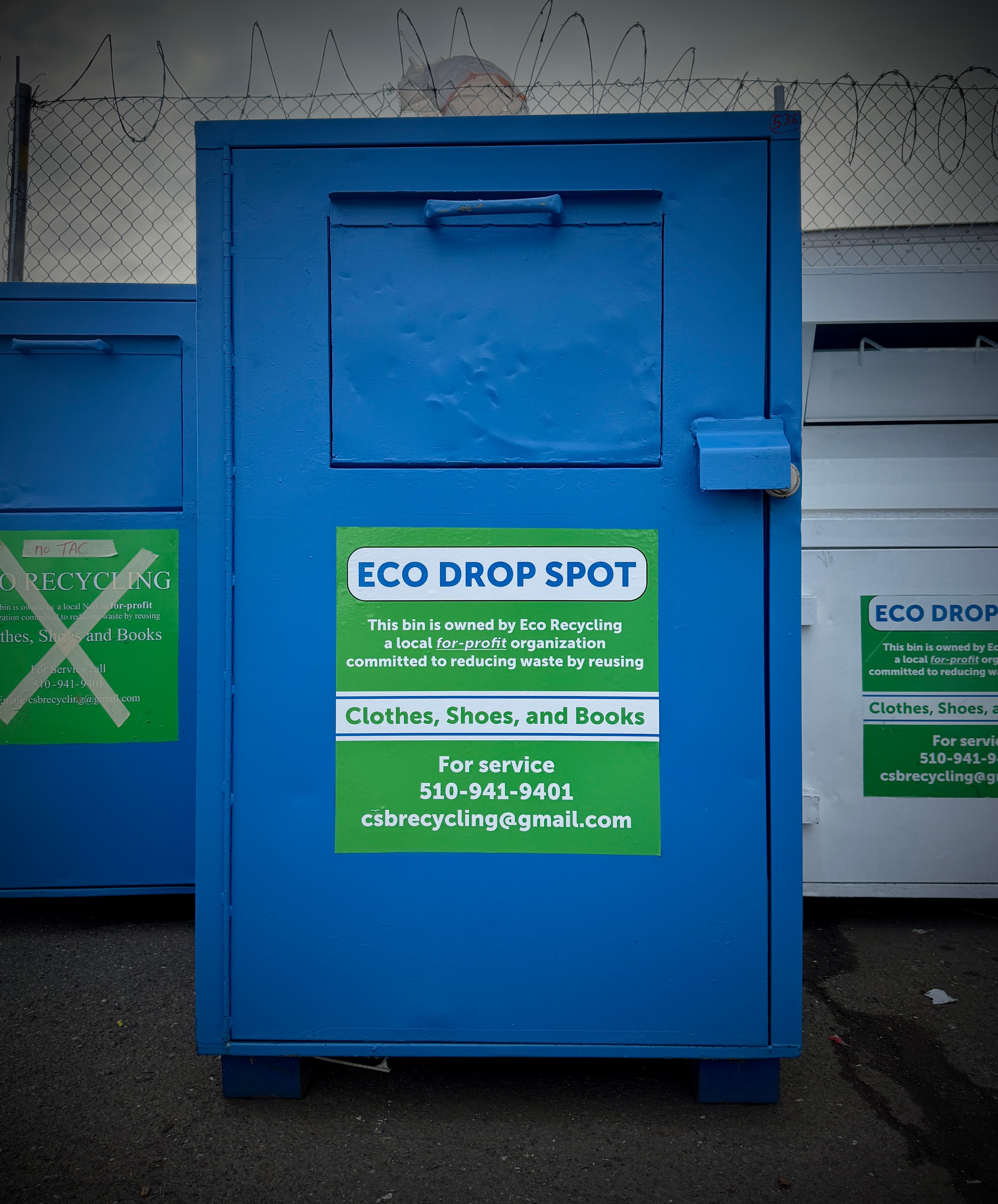 Blue Eco Drop Spot recycling bin for clothes, shoes, and books, with barbed wire fence.