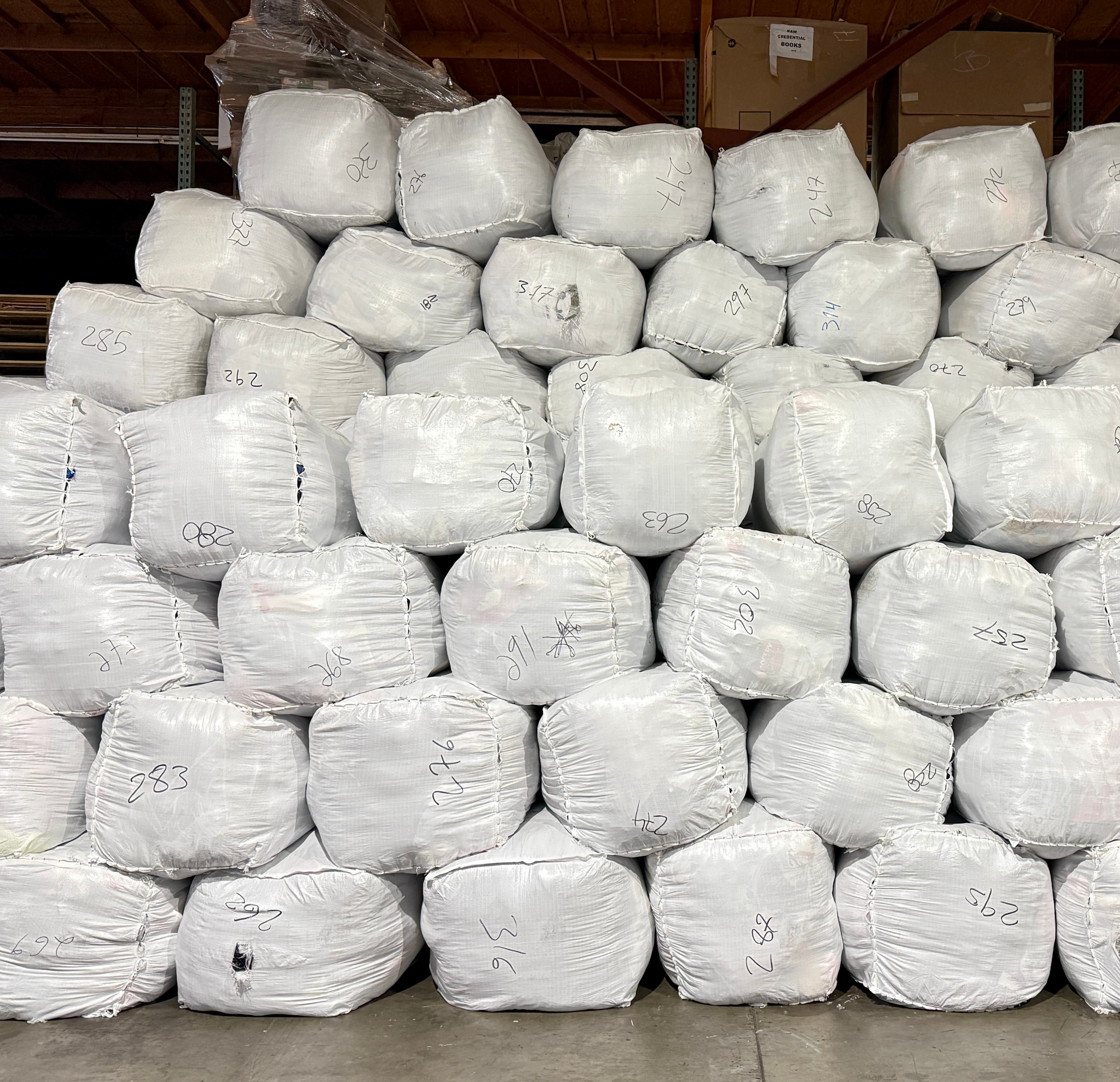 Numerous white bags with handwritten numbers stacked high in a warehouse storage area.
