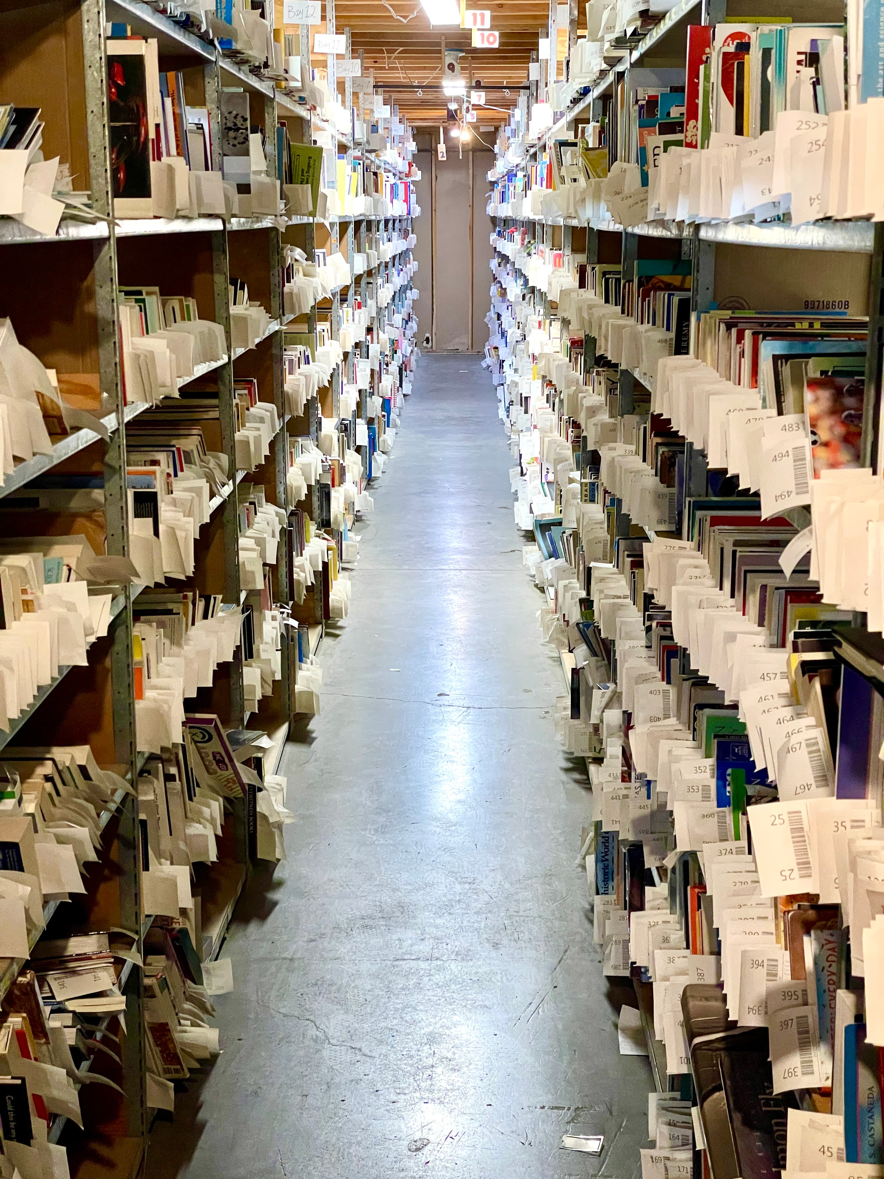 Storage aisle with metal shelves densely packed with books, each marked by white slips.